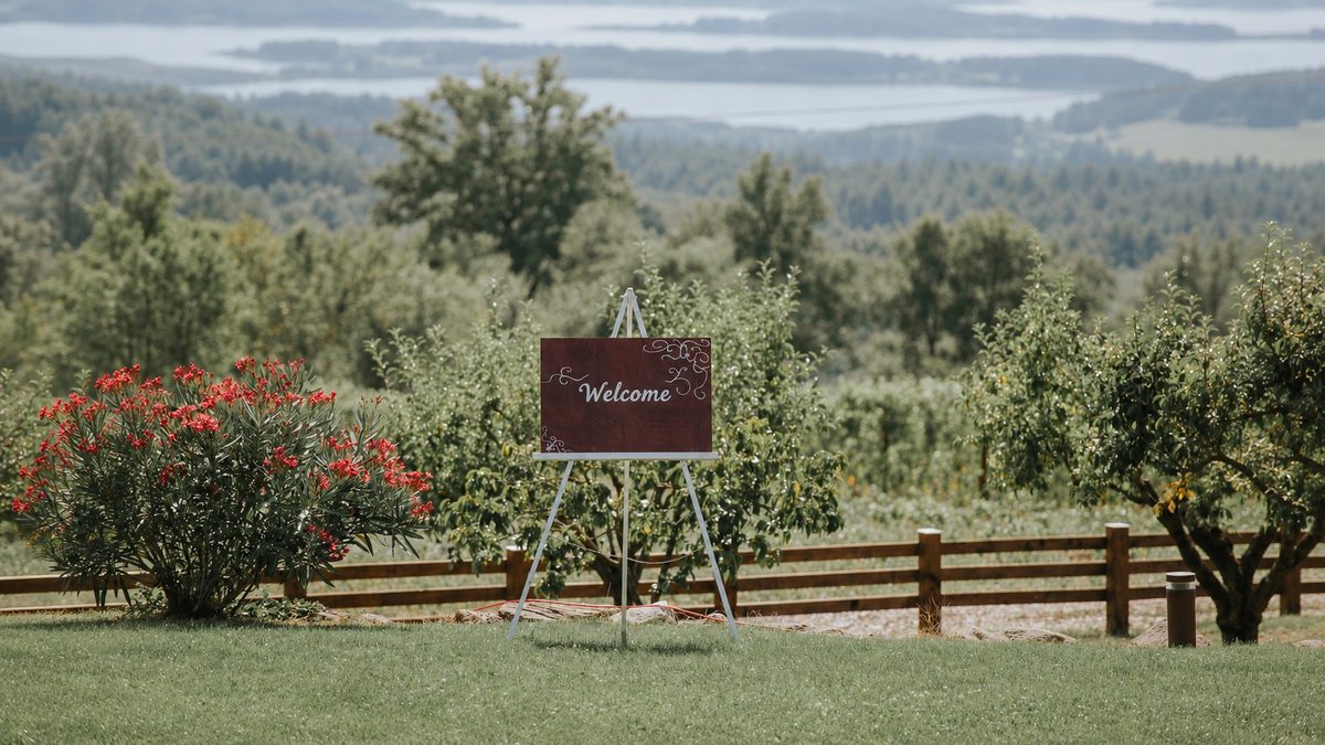 Welcome-Schild auf einer Staffelei in einem Obstgarten, dahinter Hügel und ein See